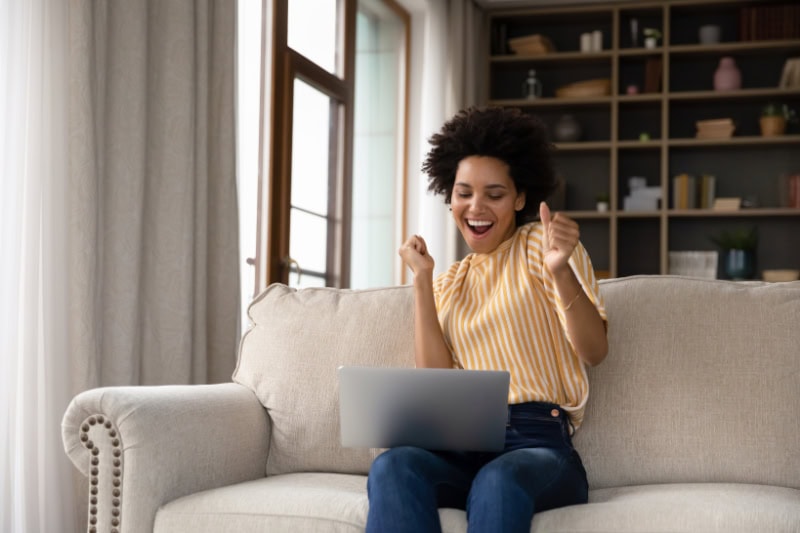 Air Conditioners: Things You May Not Have Known. Excited cheerful young Black woman using laptop computer on sofa at home, getting good news, feeling joy, dancing with hands, singing, laughing, making winner gesture, happy to win prize.