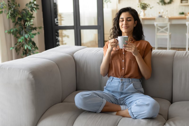 Understanding Your Air Conditioner. Young woman sitting on couch with closed eyes, drinking hot coffee at home in her living room