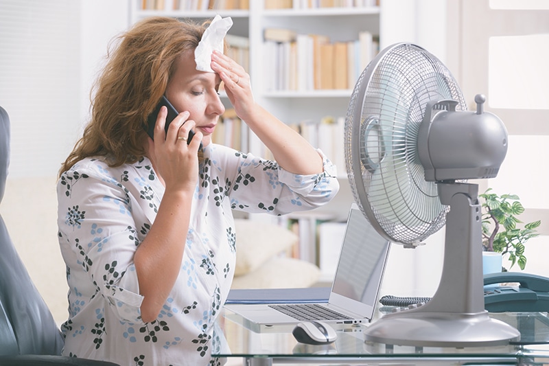 When Do I Replace My Air Conditioner? A woman sitting in front of a fan wiping sweat from her forehead.