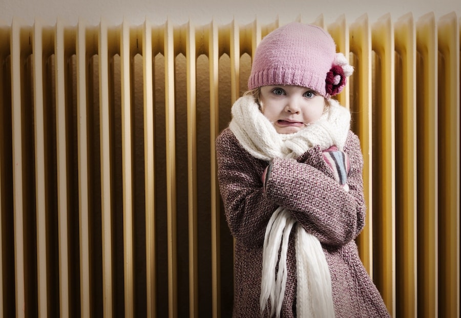 Girl in winter clothes is shivering in front of radiator. Fearful expression. Concept for energy crisis or old heating system.